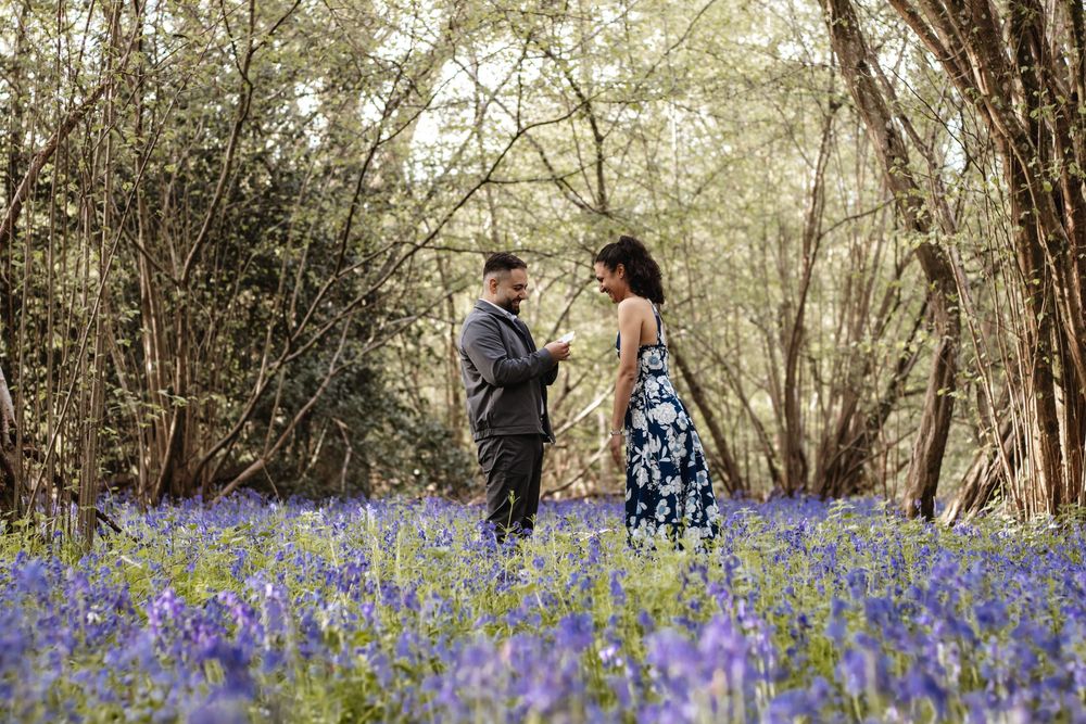 Couple standing together in a bluebell woodland forest, man in suit and woman in floral dress, romantic outdoor photo.