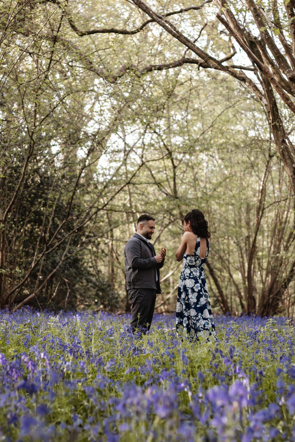 Couple standing in a bluebell woodland forest, surrounded by purple wildflowers and trees in bloom.