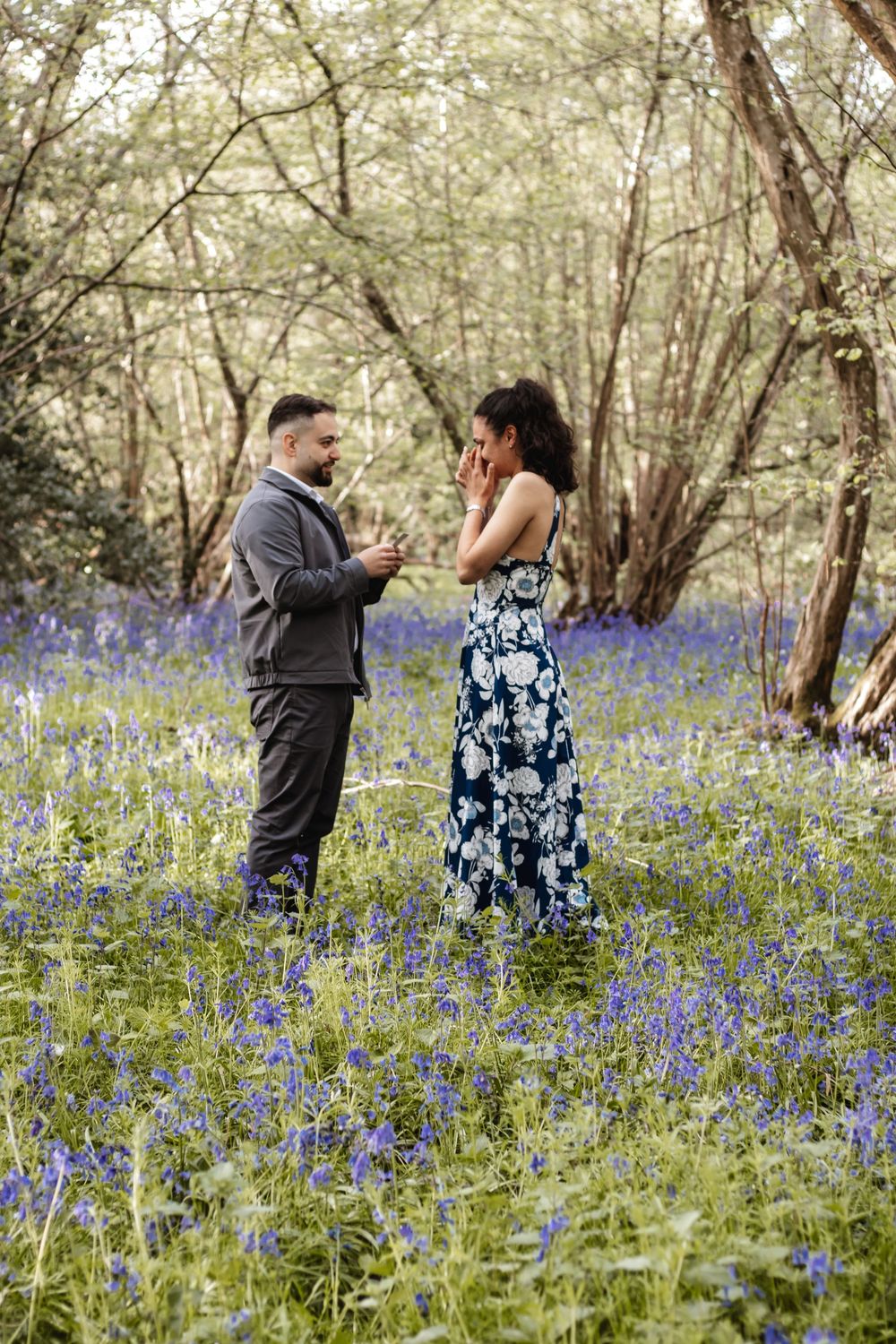 Man proposing to woman in a bluebell woodland, she covers her mouth in surprise, wearing a floral dress.