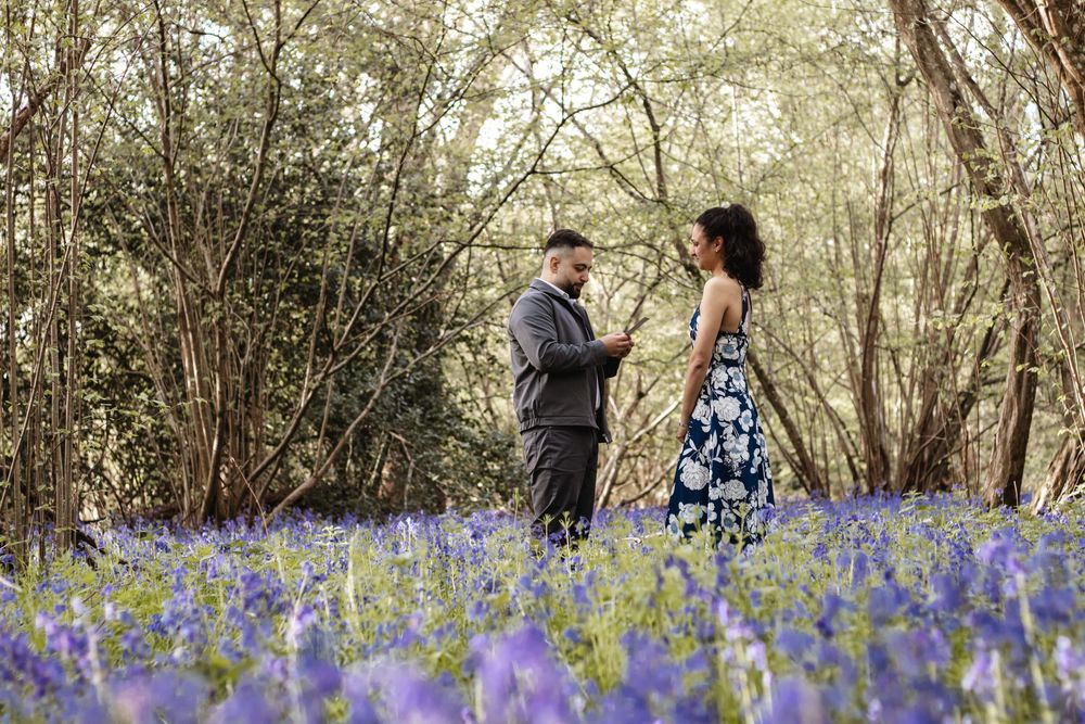 Couple standing together in a bluebell woodland, woman in floral dress and man in grey suit surrounded by purple flowers.