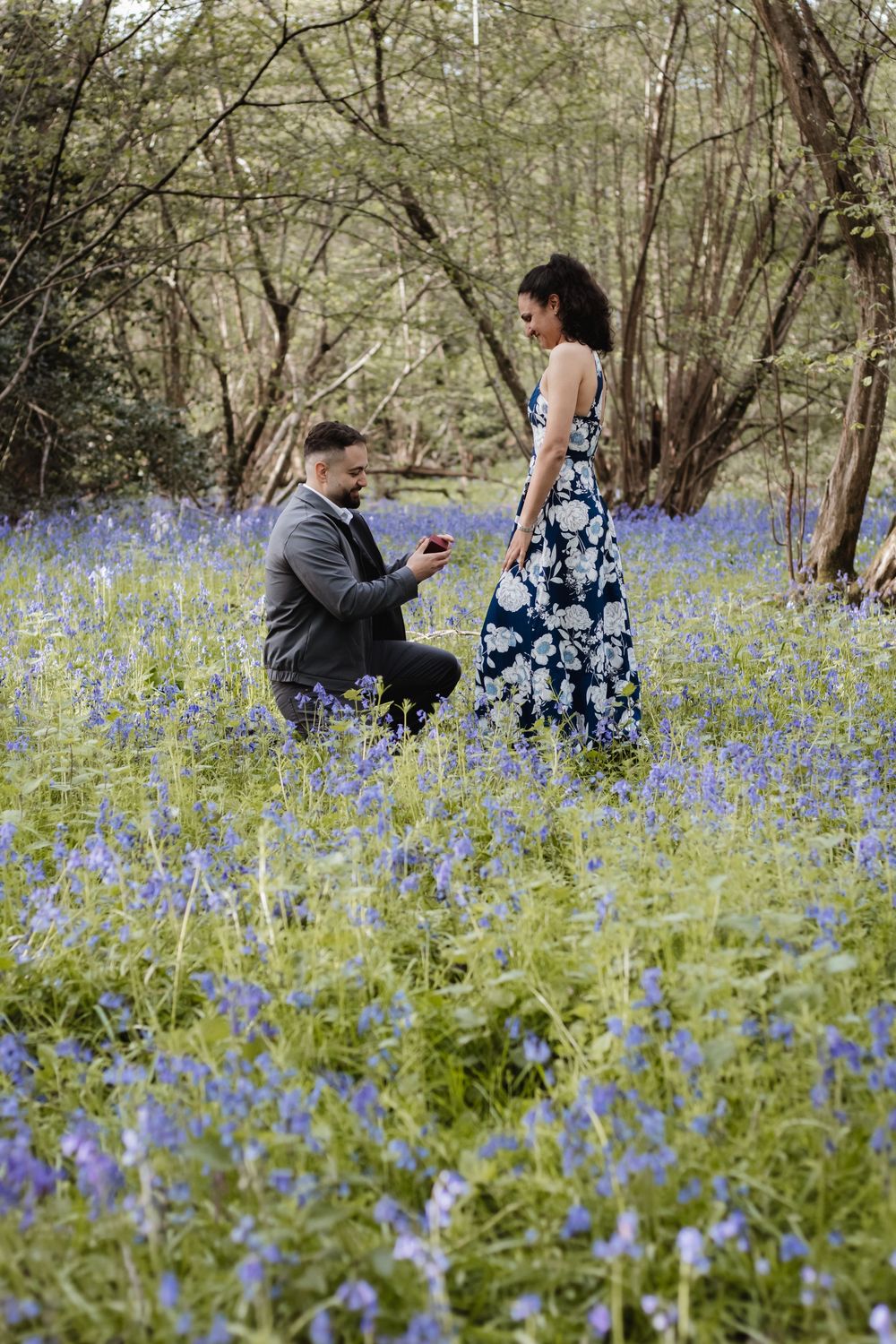 Man proposing marriage on one knee in a bluebell woodland field to woman in floral dress.