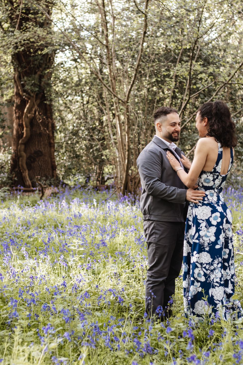 Couple embracing in a bluebell woodland, man in grey suit, woman in floral navy dress, surrounded by purple wildflowers.