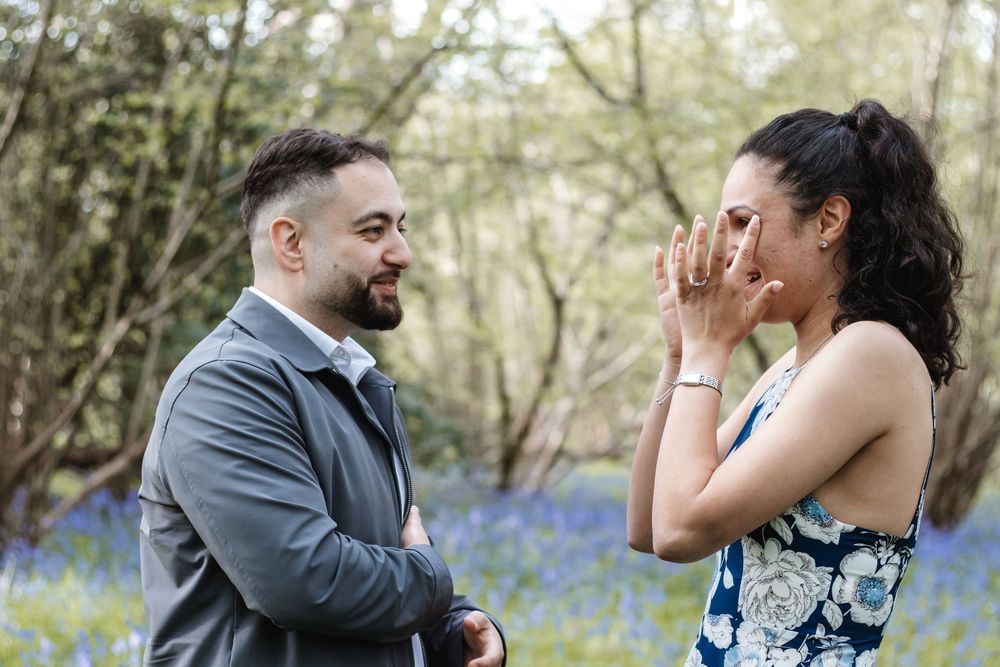 Couple sharing a romantic moment in a bluebell woodland, woman covering her mouth in surprise during a proposal.