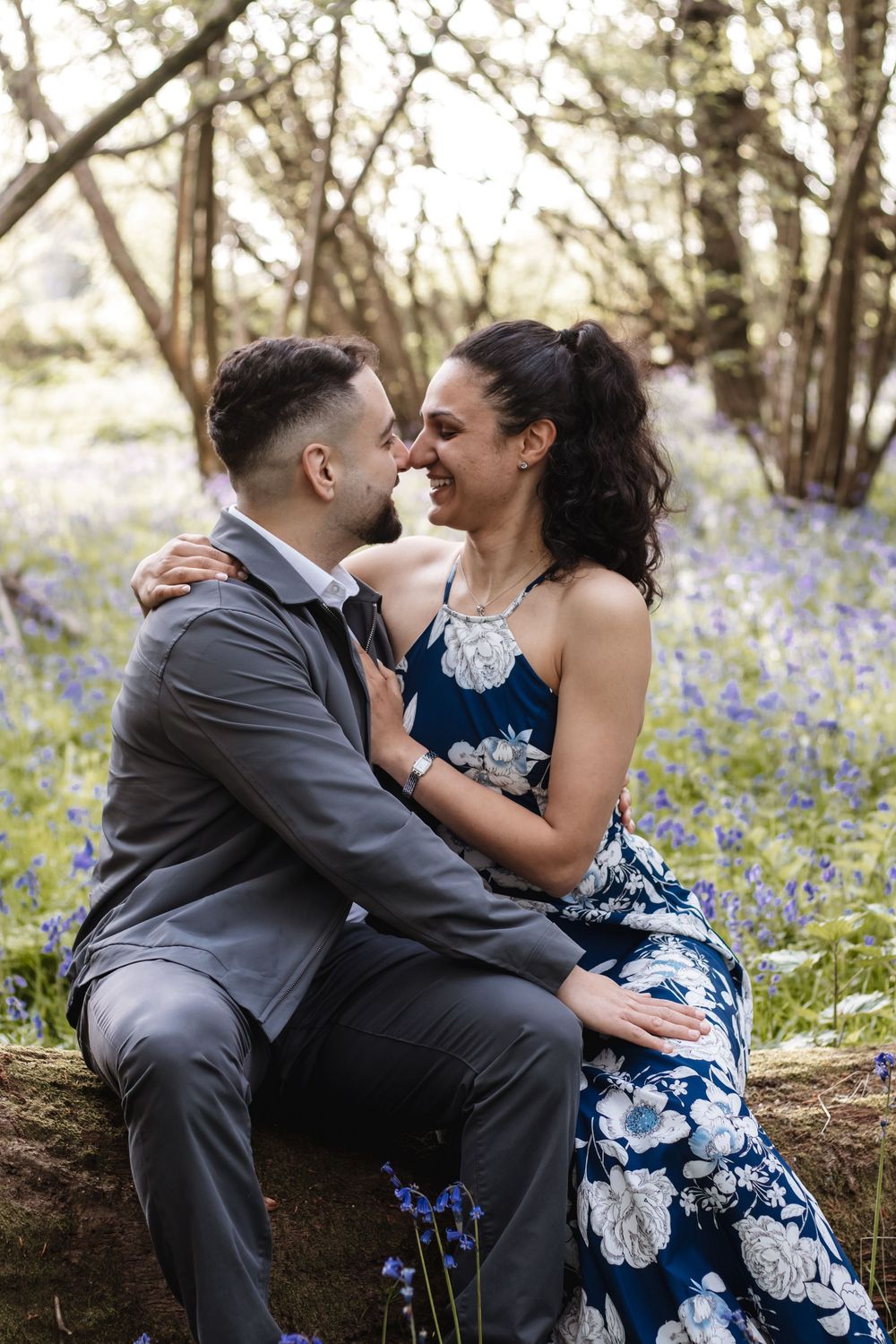Couple sharing a romantic moment sitting on a log surrounded by bluebells in a woodland setting.