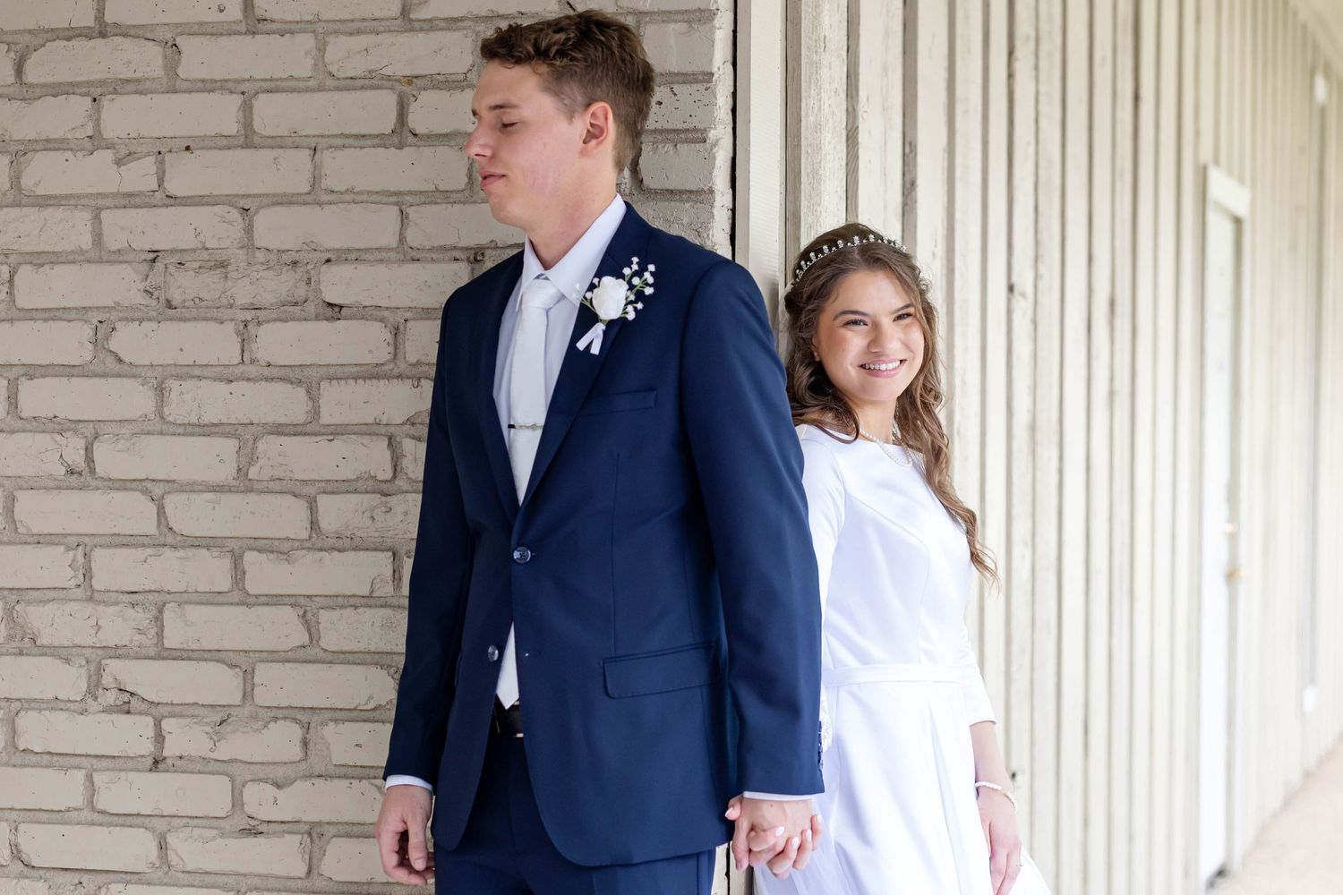 Young couple in formal wear, man in navy suit with boutonniere, woman in white dress, posing by brick wall. first touch