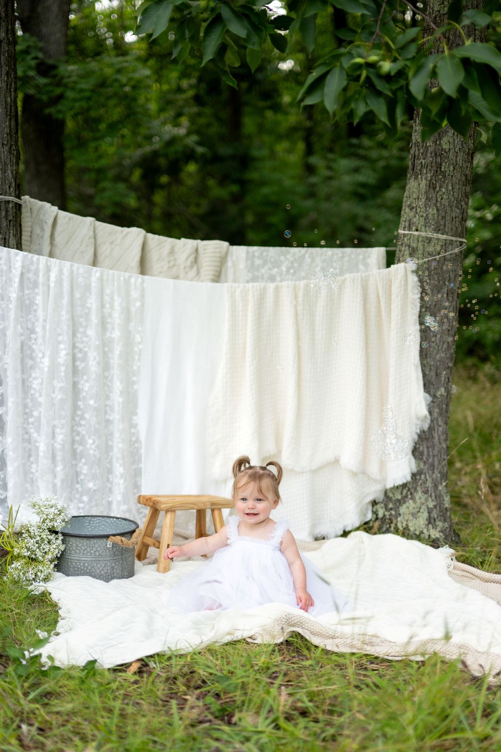 Smiling baby girl in white dress sits on blanket outdoors near clothesline with white linens hanging between trees.