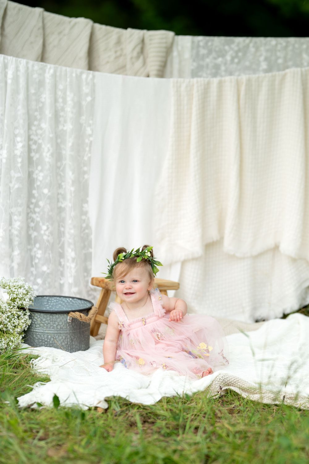 Smiling baby girl in pink dress and floral crown sits on white blanket outdoors near vintage bucket and clothesline.