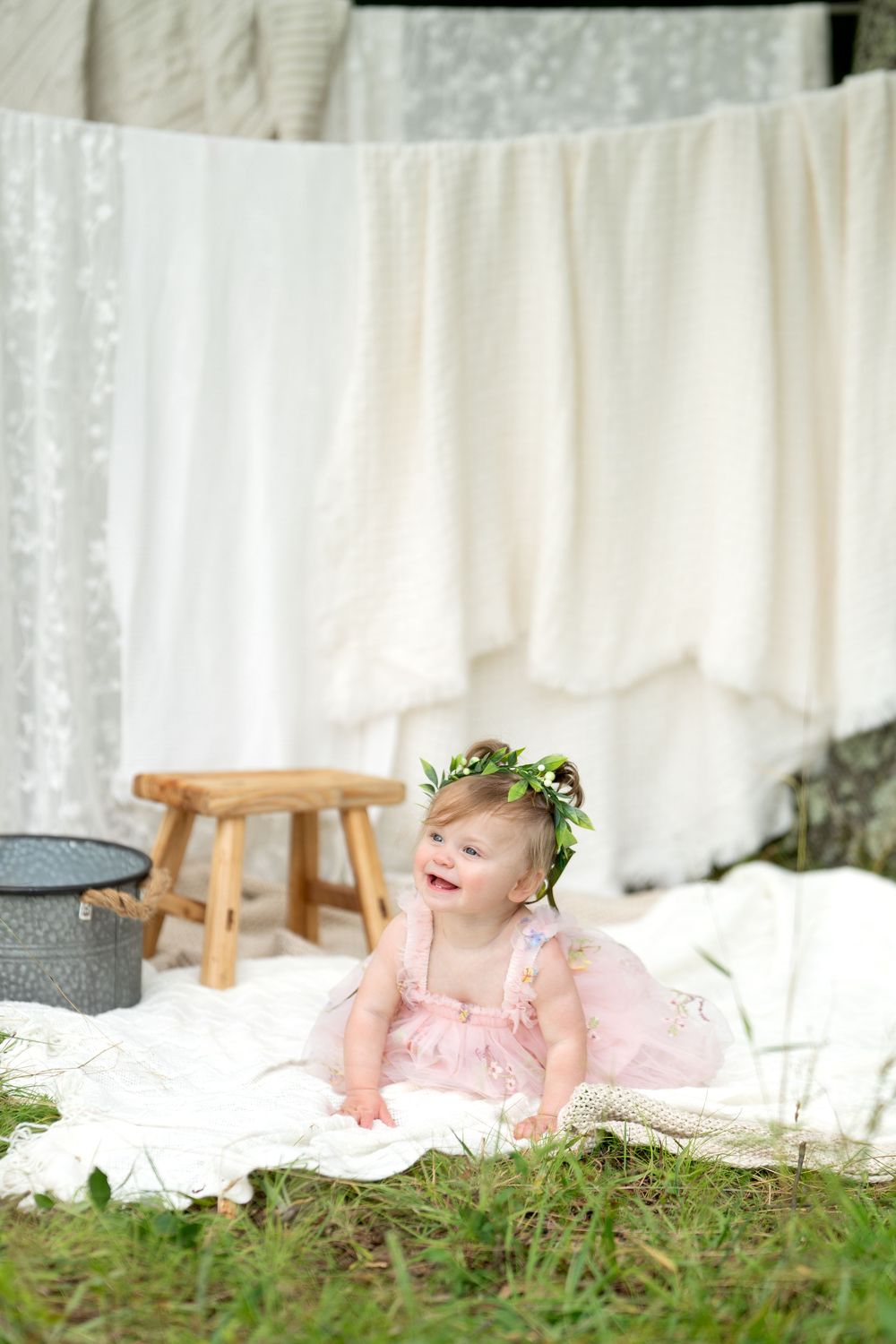 Adorable baby girl in pink dress and floral crown sitting on white blanket outdoors near hanging laundry.
