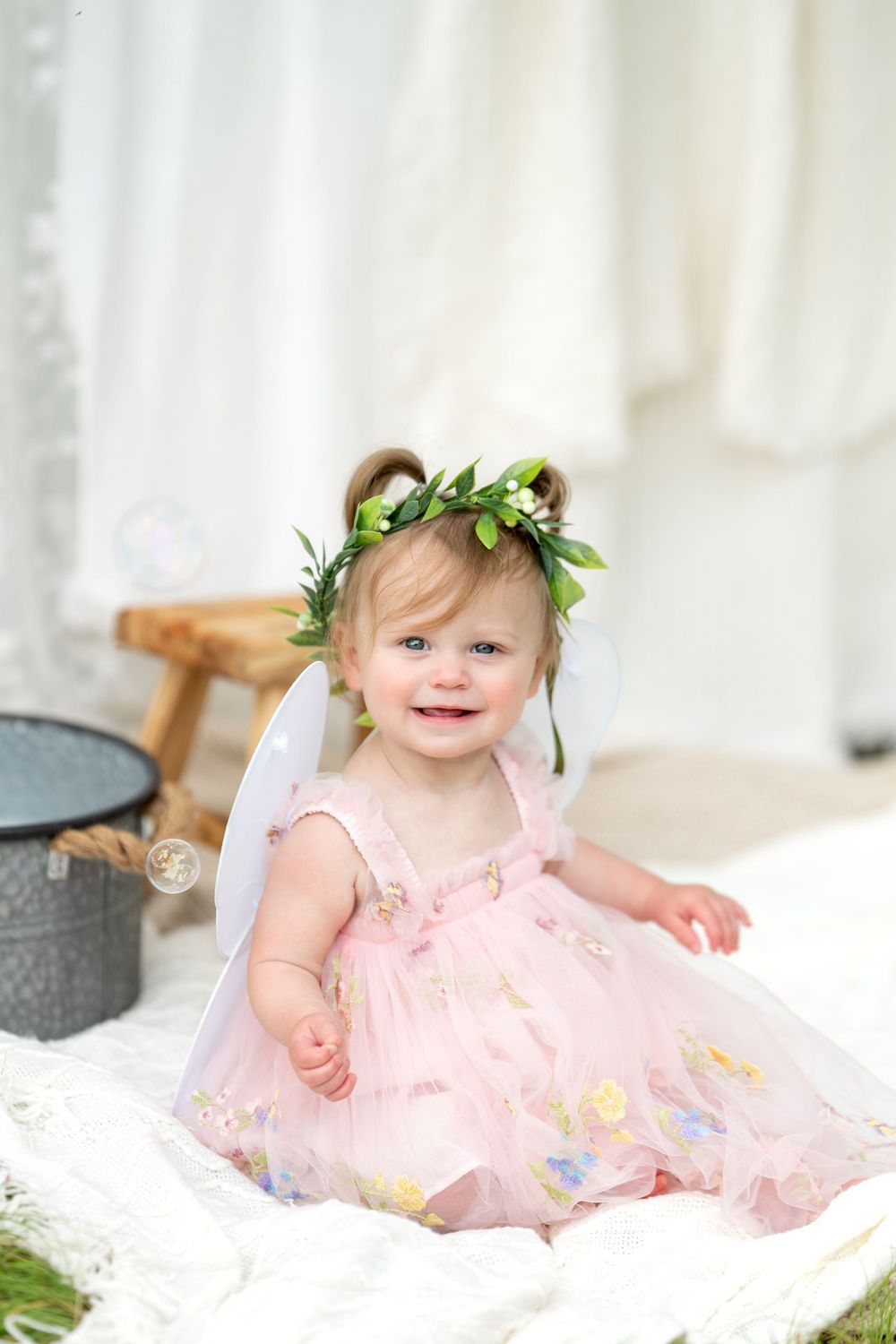 Adorable baby girl wearing pink dress, green leaf crown, and white fairy wings smiles while sitting outdoors.