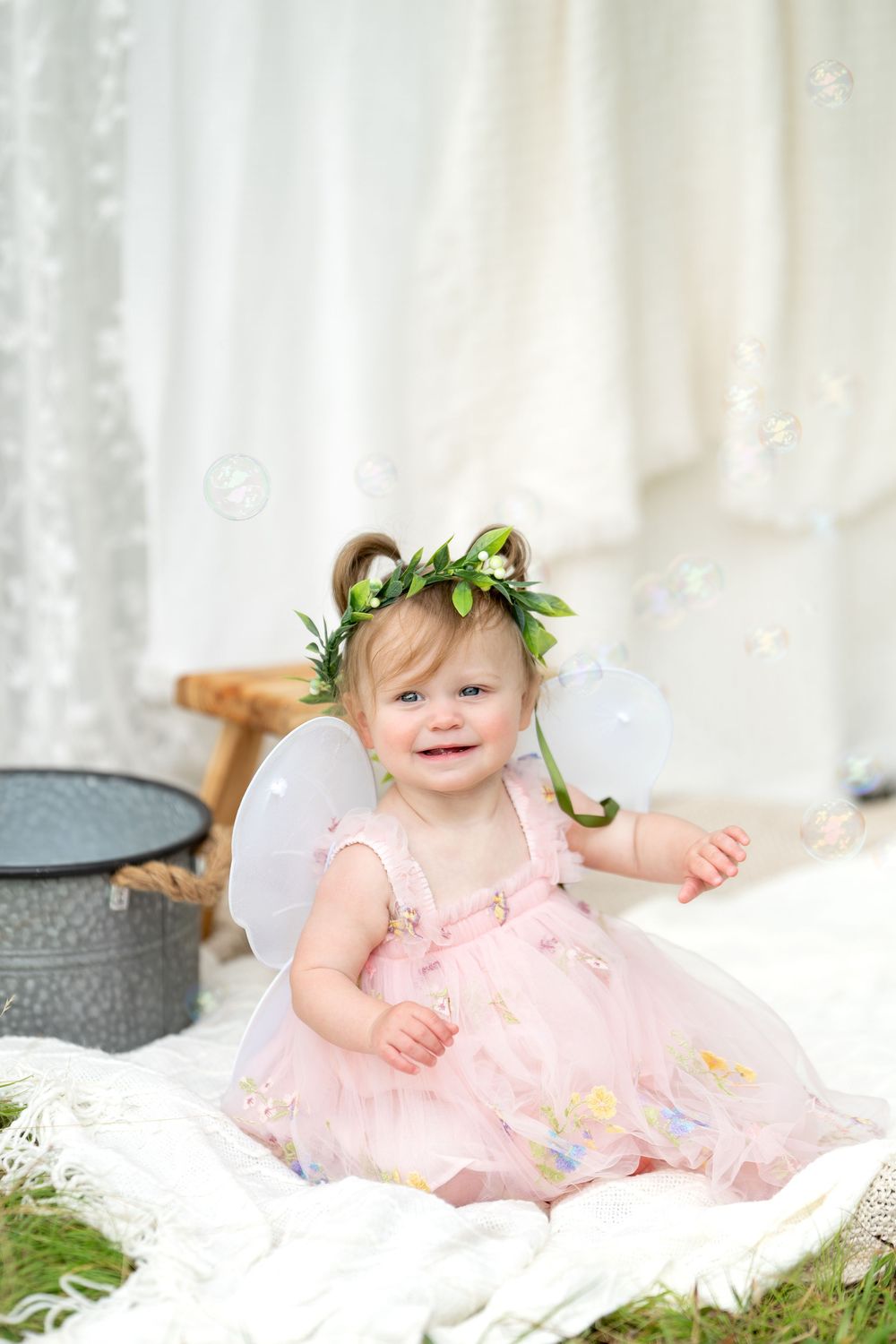 Smiling toddler girl in pink fairy dress with white wings and floral crown sits on white blanket surrounded by bubbles.