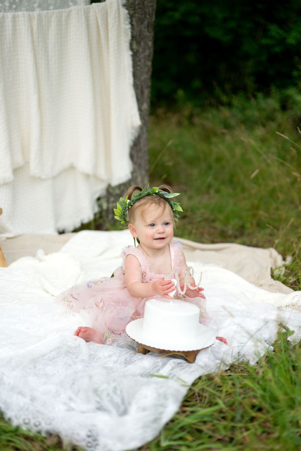 Baby girl in pink tutu dress and floral crown sits on white lace blanket outdoors near a white birthday cake.