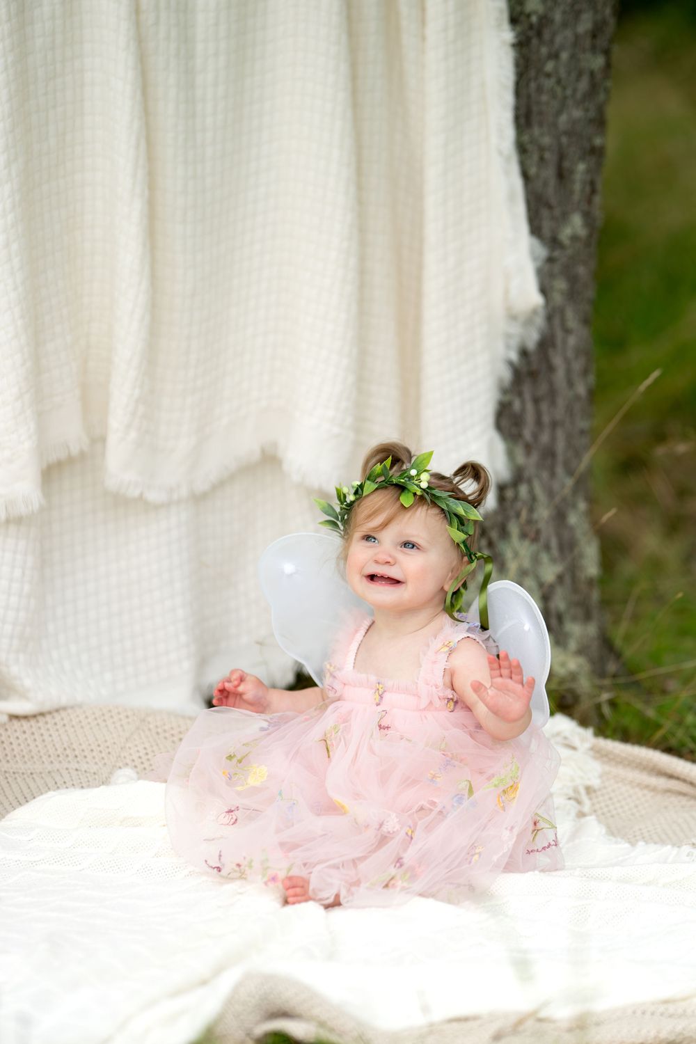Smiling baby girl in pink fairy dress with white wings and floral crown sitting on white blanket outdoors.