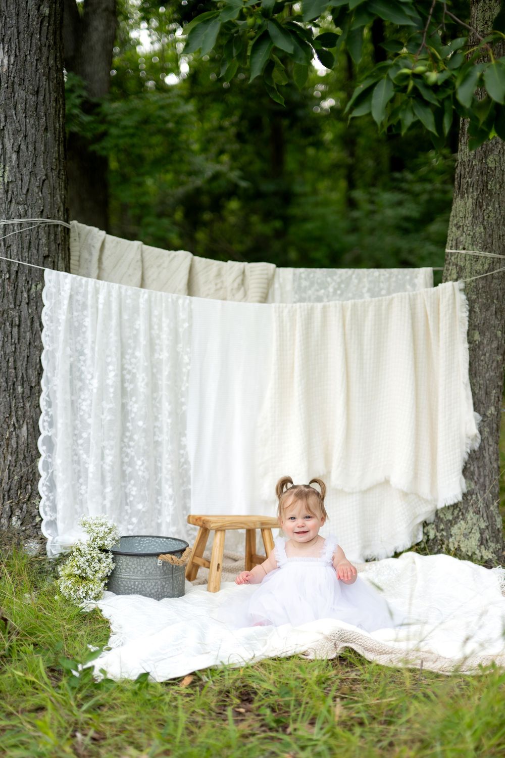 Smiling baby girl in white dress sits on blanket outdoors near clothesline with white linens hanging between trees.