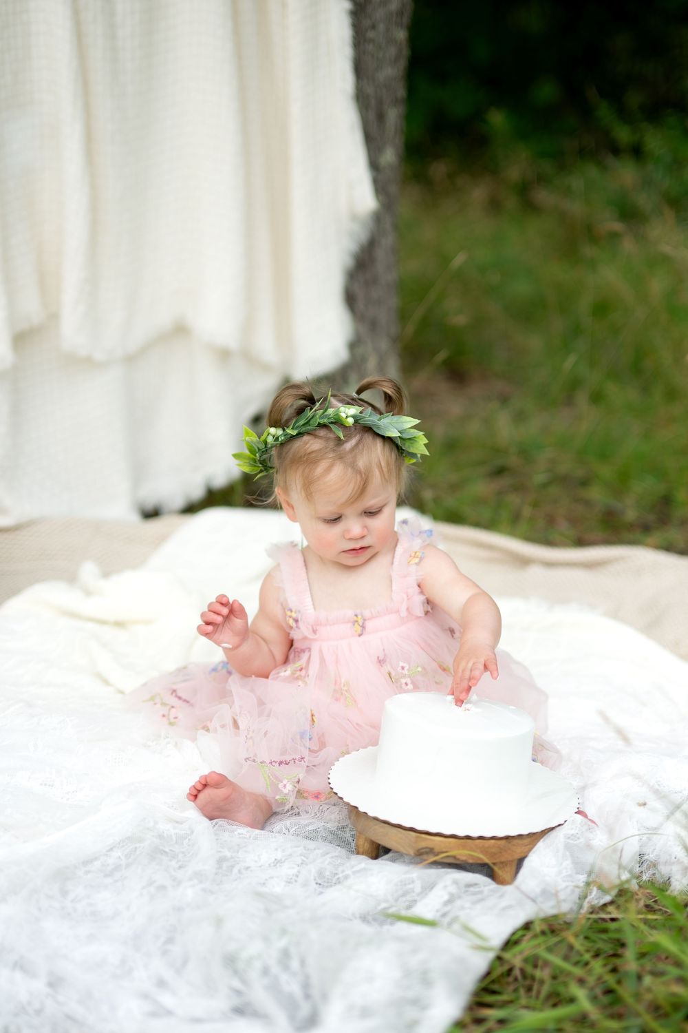 Adorable baby girl in pink dress with floral crown reaches toward white smash cake on wooden stand outdoors.