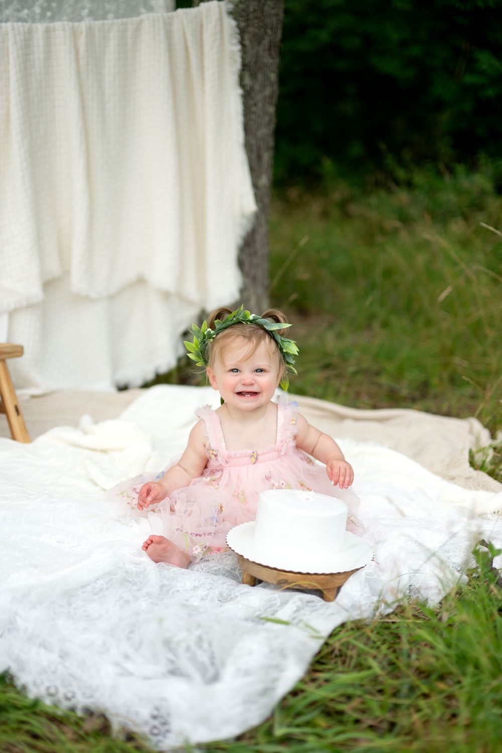 Smiling baby girl in pink dress and floral crown sits on white blanket outdoors near tree for first birthday cake smash.