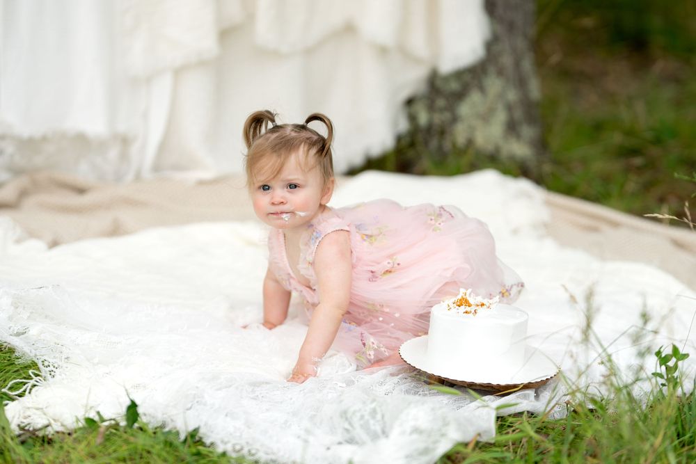 Baby girl in pink tutu dress crawling on white blanket outdoors near a small white smash cake.