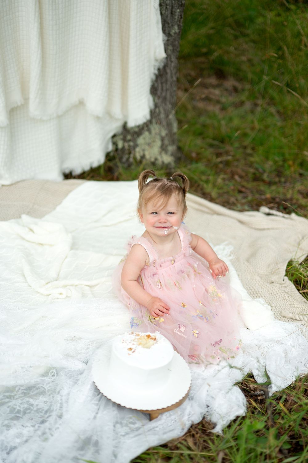 Smiling baby girl in pink floral dress sits on white blanket outdoors next to her smashed first birthday cake.