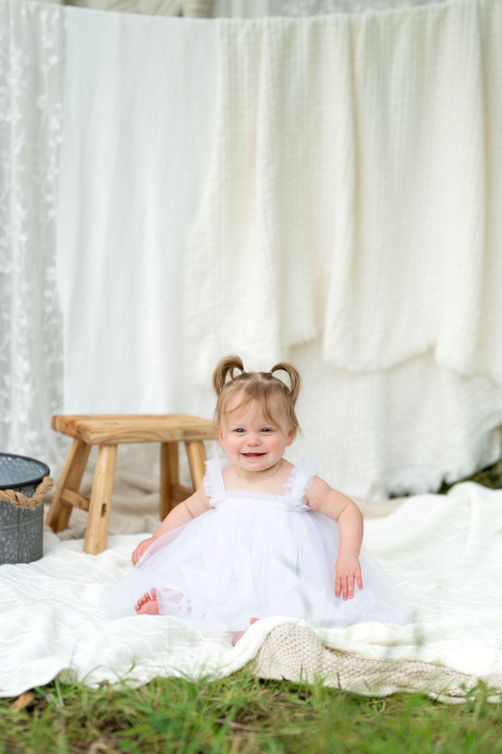 Smiling toddler girl in white dress sitting on blanket outdoors with white linens hanging in background.