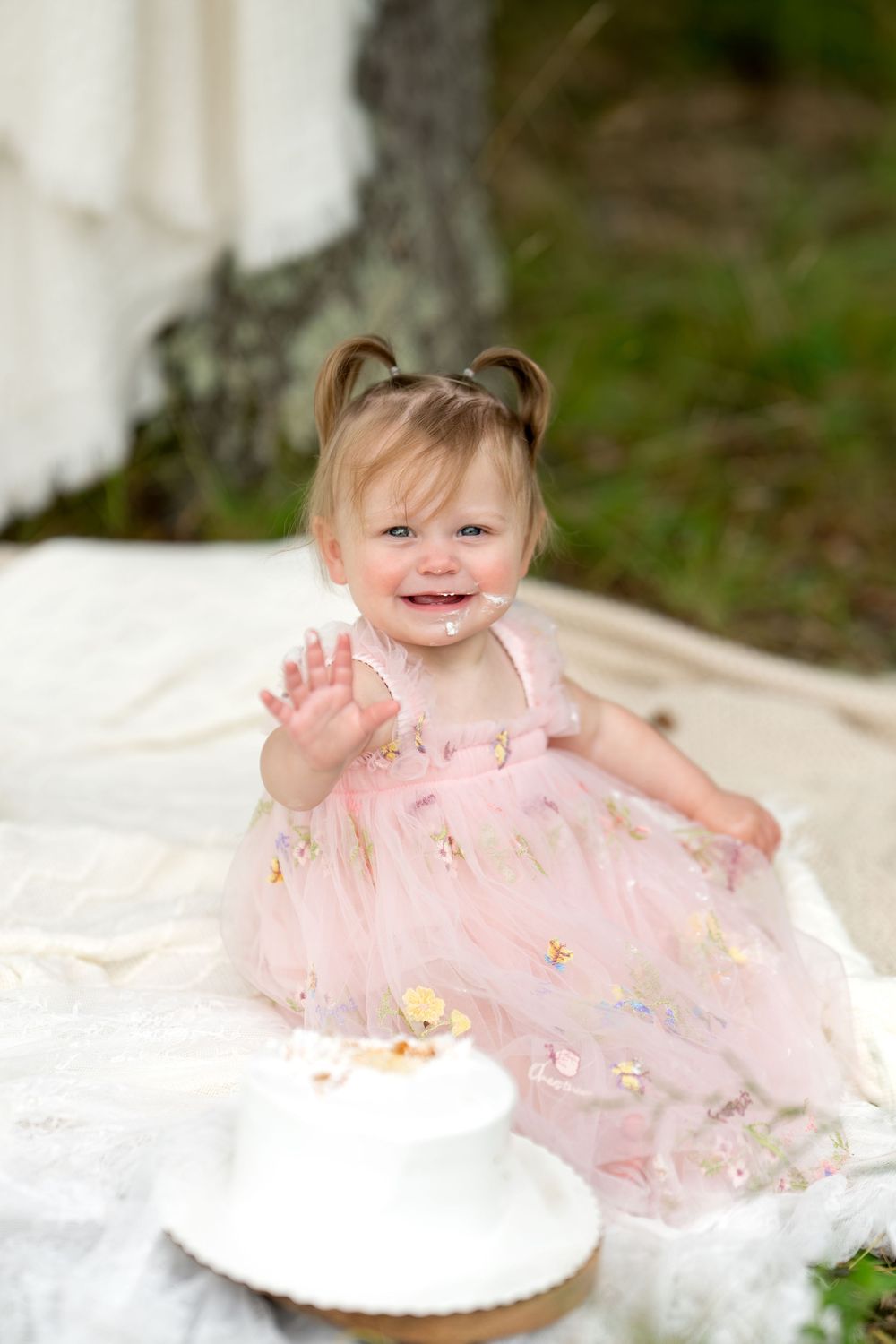 Happy baby girl in pink dress smiling with cake on face, waving at first birthday.