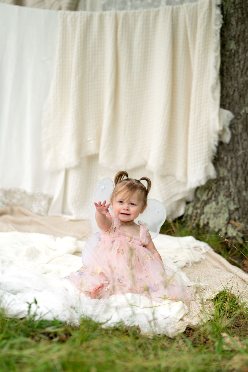 Baby girl in pink floral dress and white fairy wings waves hand while sitting on blanket outdoors.