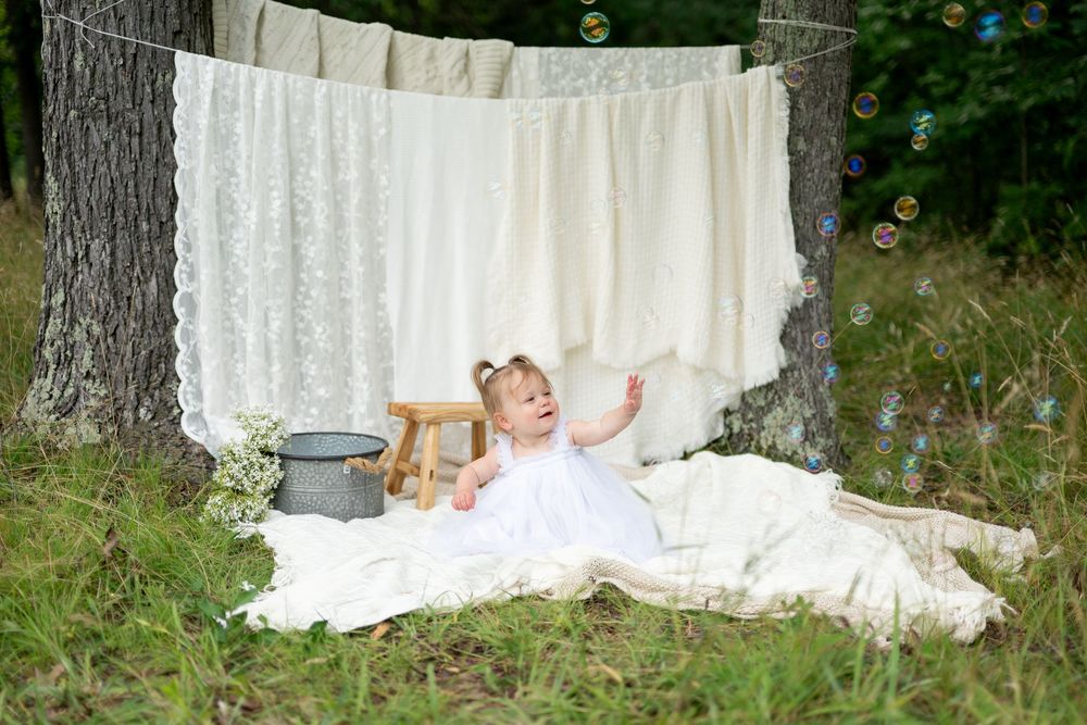 Baby girl in white dress sits on blanket outdoors near hanging lace curtains with bubbles floating nearby.