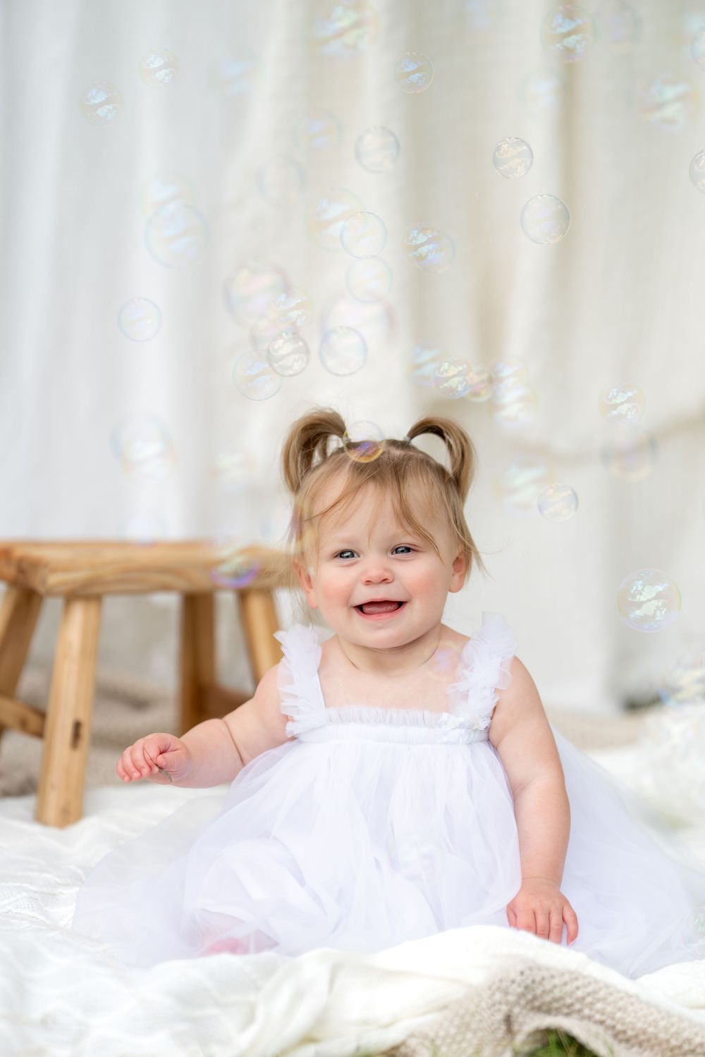 Smiling baby girl in white dress surrounded by floating soap bubbles during a studio photo session.
