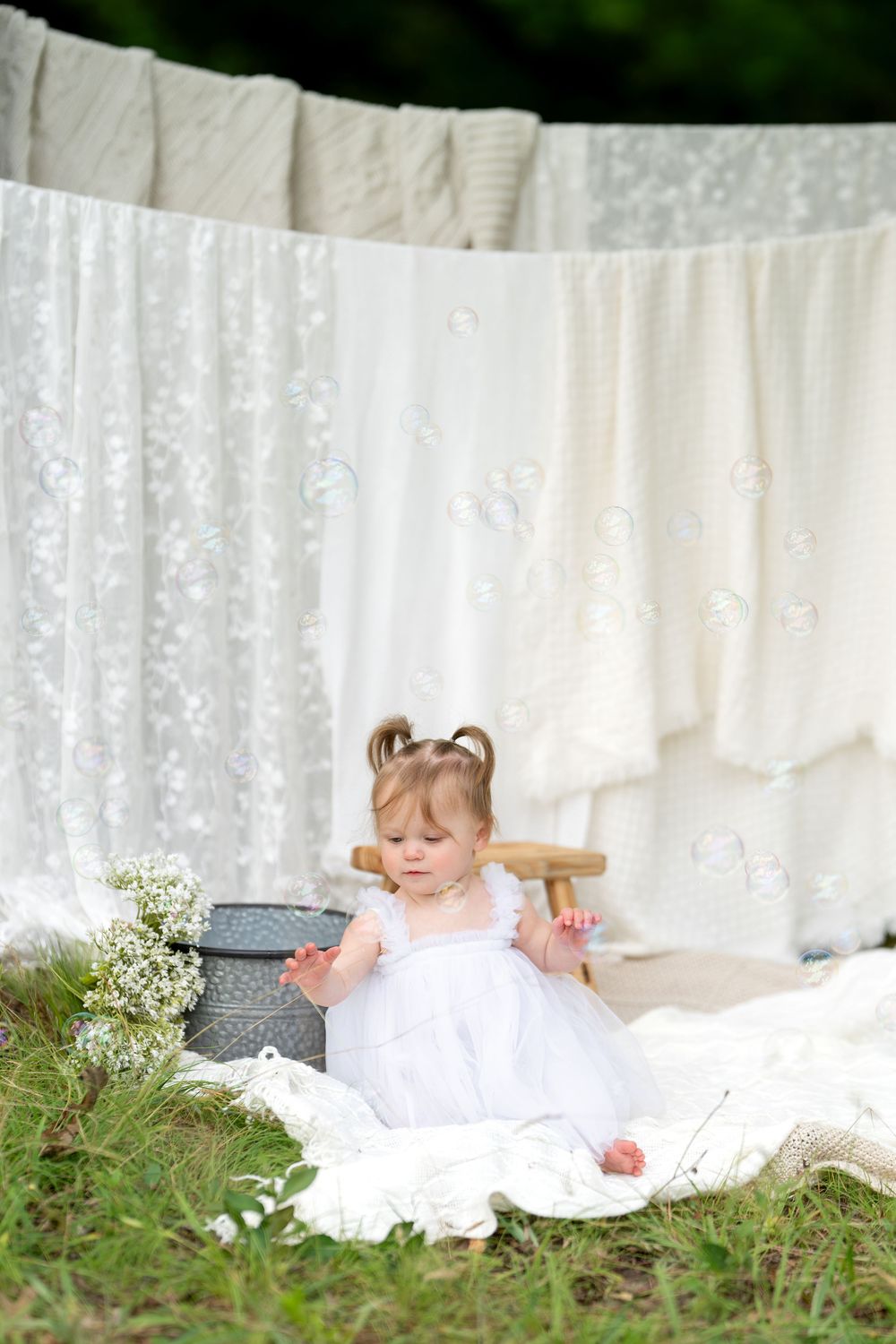 Cute toddler girl in white dress plays with wooden stool near clothesline with white linens hanging outdoors.
