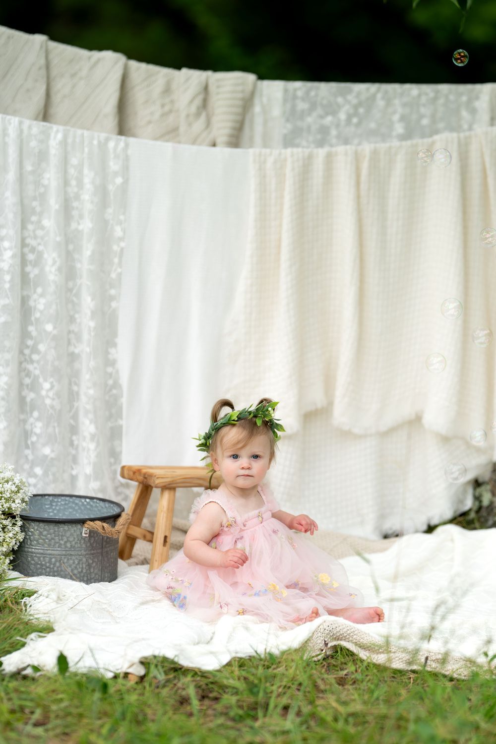 Baby girl in pink floral dress and green leaf crown sits on white blanket outdoors near hanging laundry.