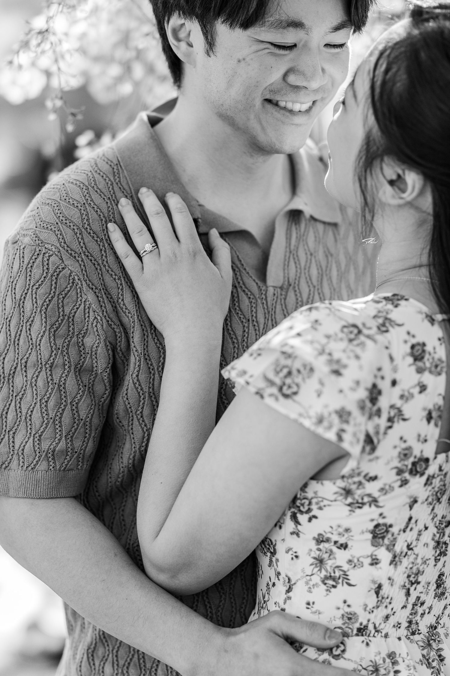 Black and white photo of a smiling couple embracing, woman wearing engagement ring resting hand on man's chest.
