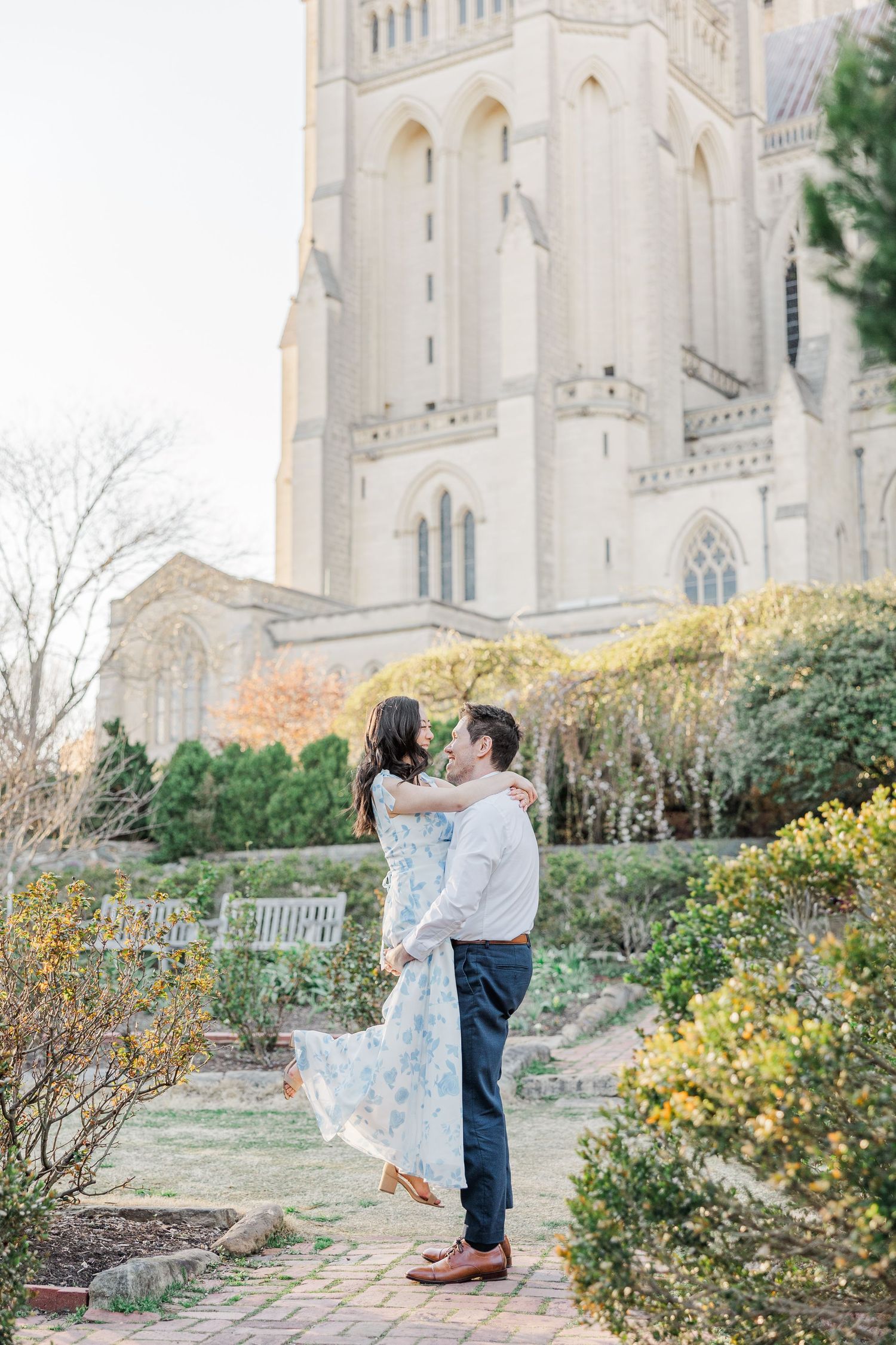 Couple embracing in a garden outside a Gothic cathedral, woman in floral dress lifted by man in white shirt.