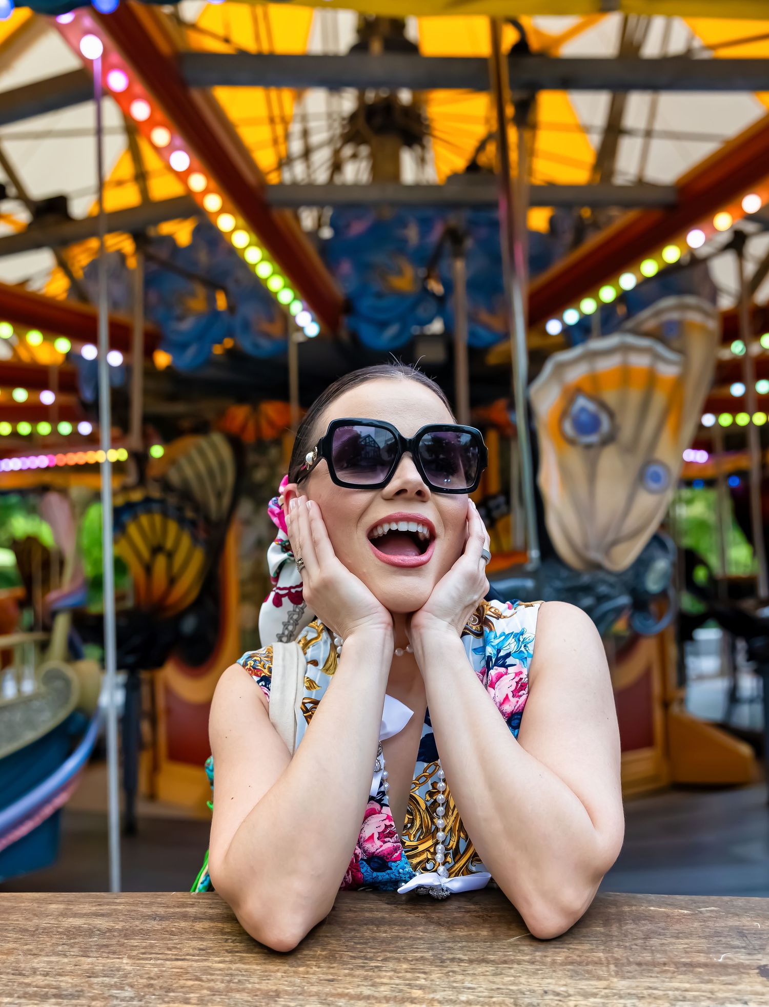 Excited woman in sunglasses and floral top poses at a colorful carousel, hands on cheeks, mouth open in joy.