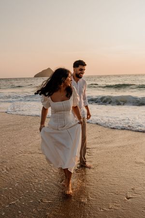 Couple in white outfits walking barefoot on a sandy beach at sunset with ocean waves.