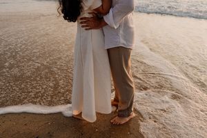 Couple embracing barefoot on sandy beach shore at sunset, waves washing around their feet.