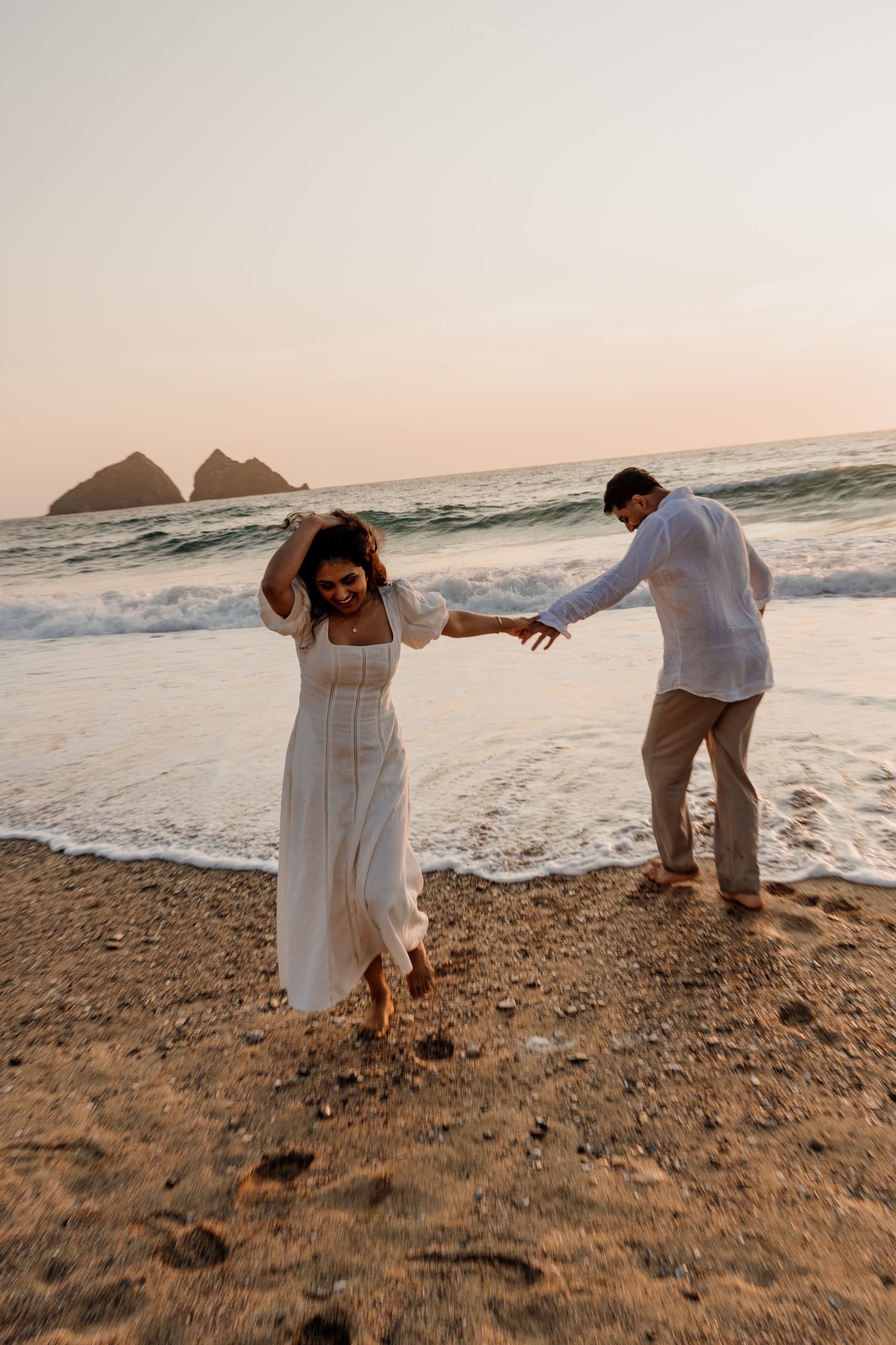 Couple dancing playfully on a sandy beach at sunset, woman in white dress, ocean waves in background.