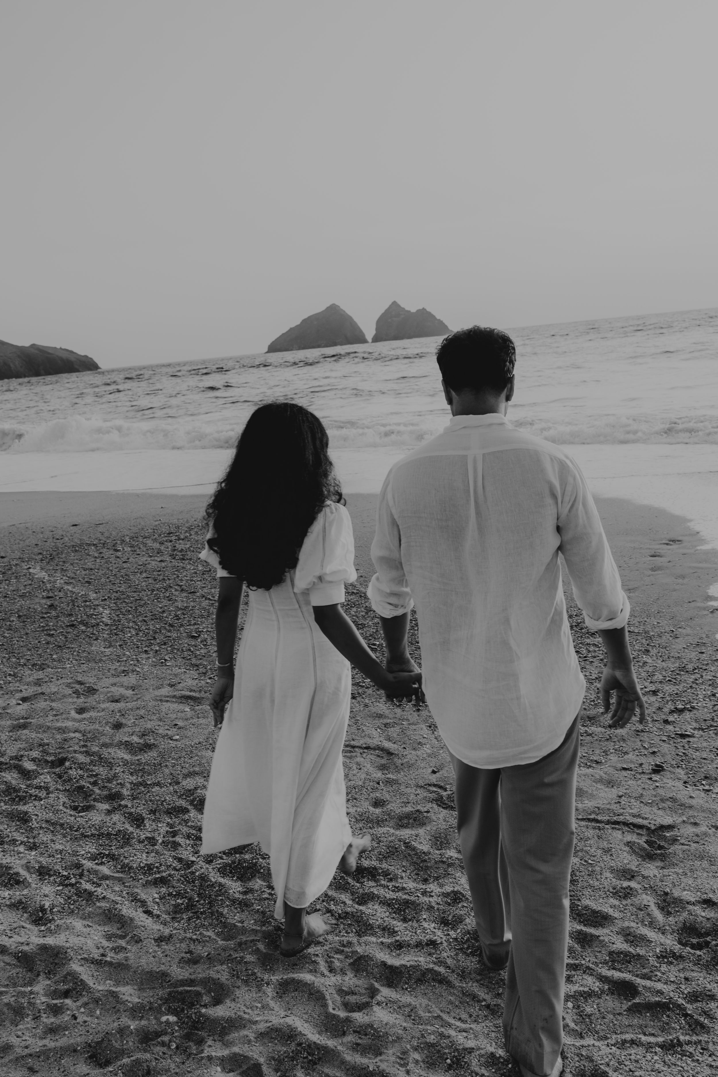 Black and white photo of a couple holding hands walking on a sandy beach toward rocky ocean formations.