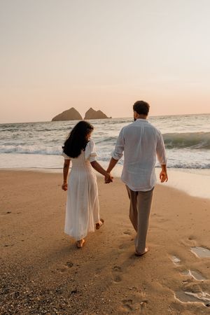 Couple holding hands walking barefoot on sandy beach at sunset, woman in white dress, man in white shirt.