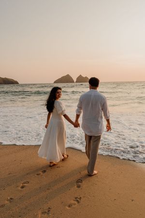 Couple holding hands walking on sandy beach at sunset wearing white outfits with ocean waves and rocky islands in background.