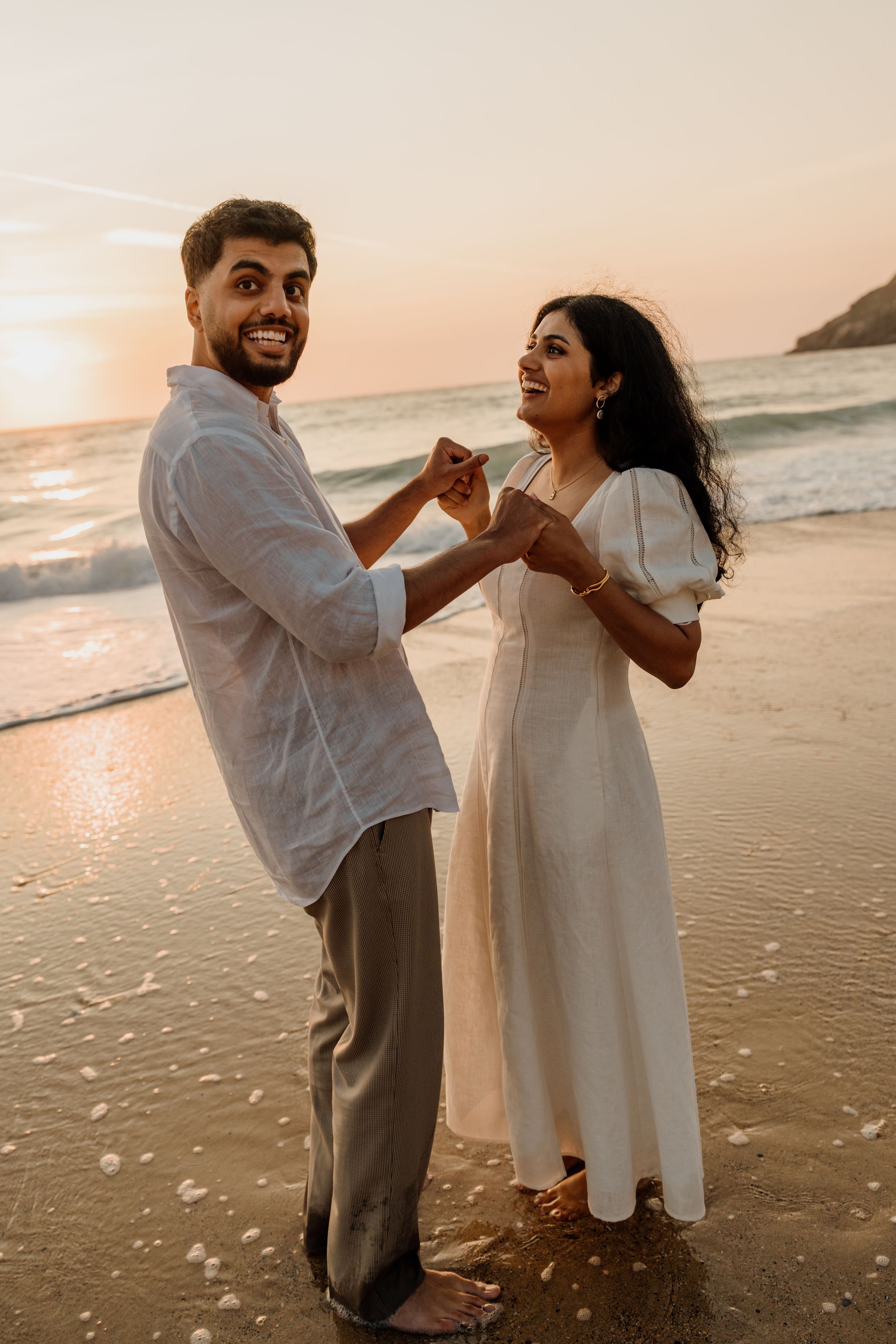 Couple holding hands and dancing barefoot on a beach at golden sunset, wearing white outfits.