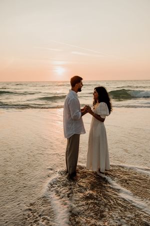 Couple in white outfits holding hands at ocean's edge during a beautiful golden sunset.