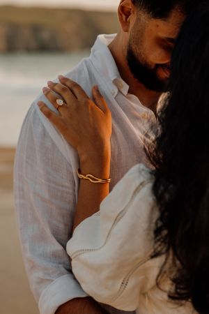 Close-up of couple embracing on beach, woman wearing diamond engagement ring and gold bracelet.