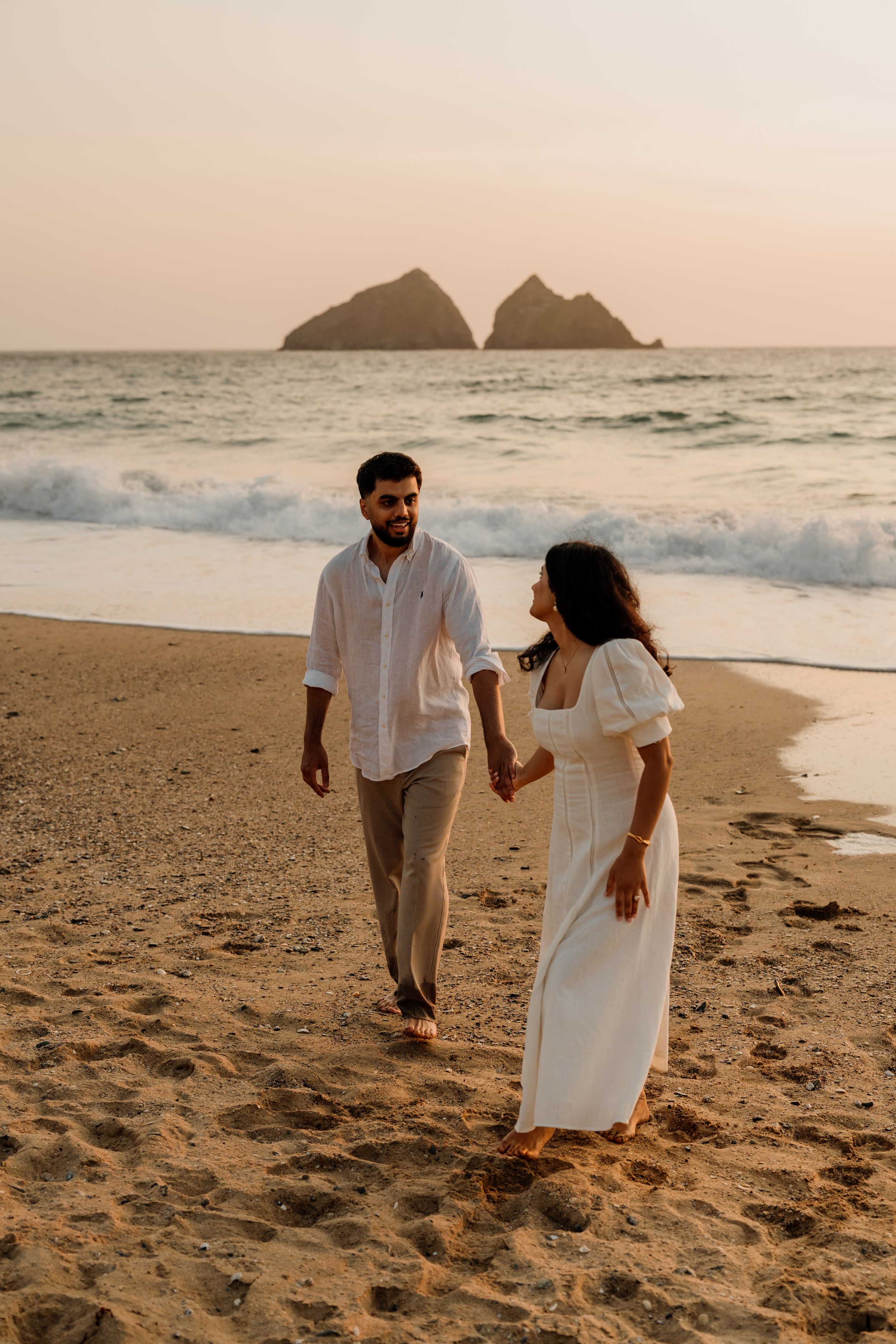 Couple holding hands on a sandy beach at sunset with twin rock islands in the background.