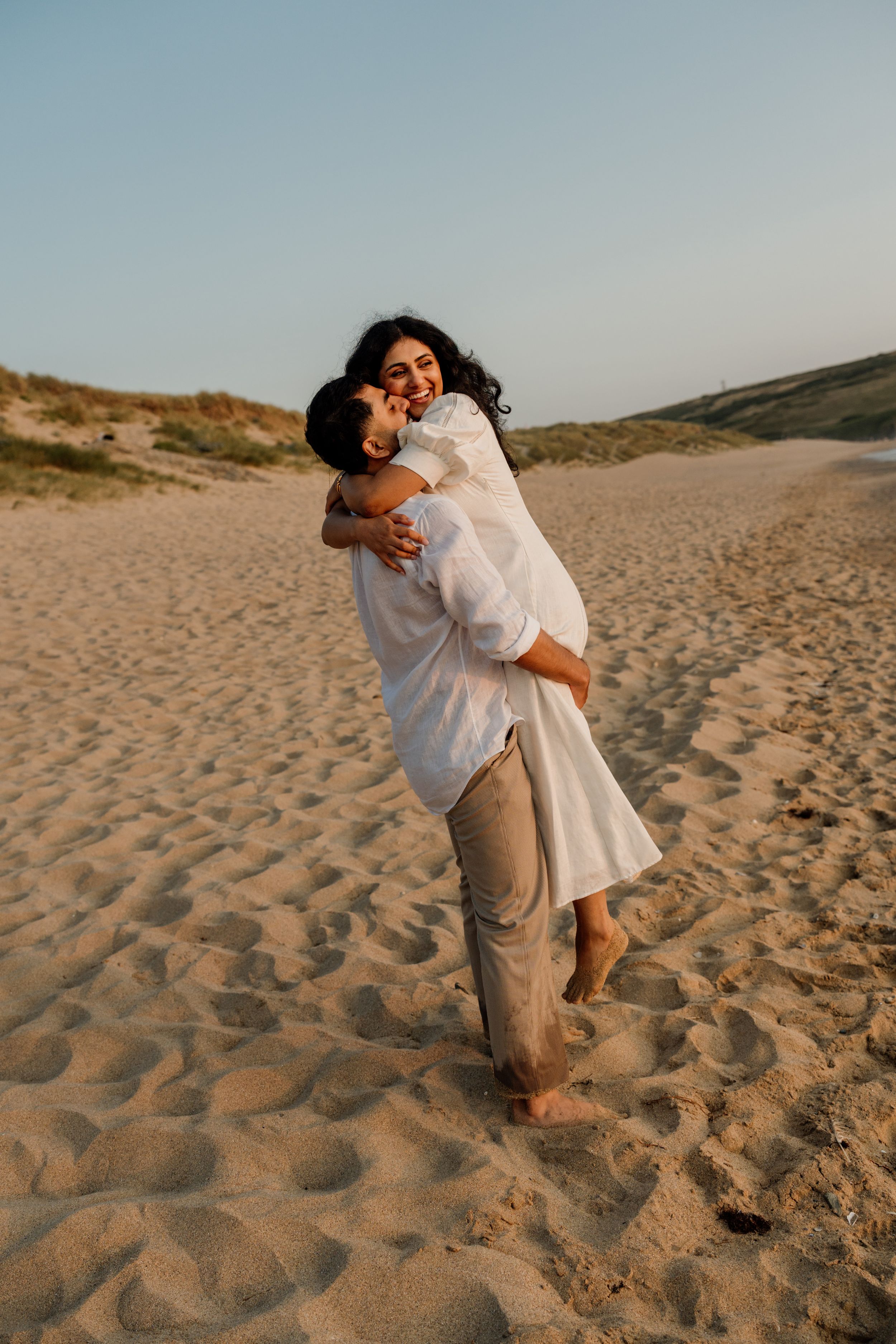 Couple embracing on sandy beach dunes at sunset, woman in white dress laughing as man lifts her.