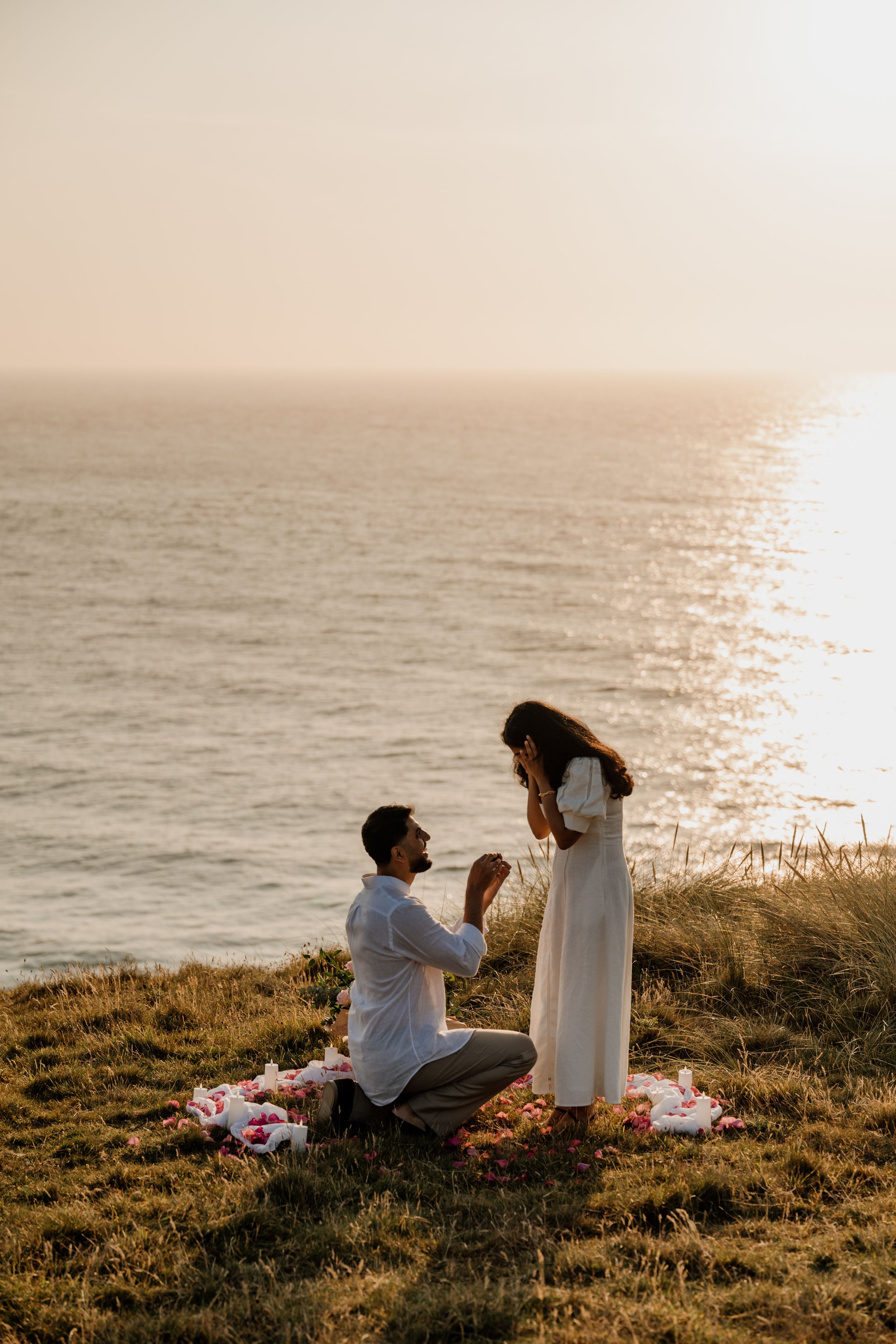 Man proposing to woman on a cliffside at sunset with ocean views and flower petals.