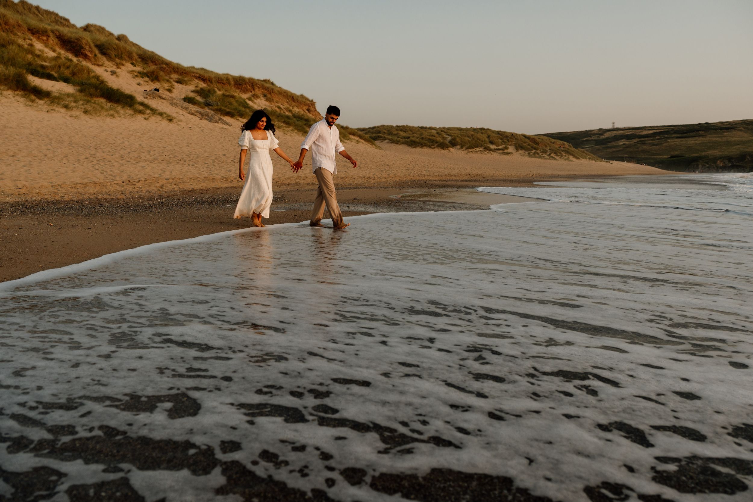 Couple walking hand in hand along a sandy beach shore at sunset, waves gently washing over their feet.