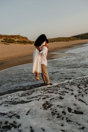Romantic couple hugging at ocean shoreline at dusk, waves washing around their feet on sandy beach.