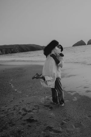 Black and white photo of a couple embracing on a sandy beach at dusk with ocean waves and rocky cliffs in background.
