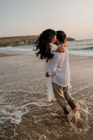 Couple sharing a kiss while man lifts woman in ocean waves on a sandy beach at sunset.