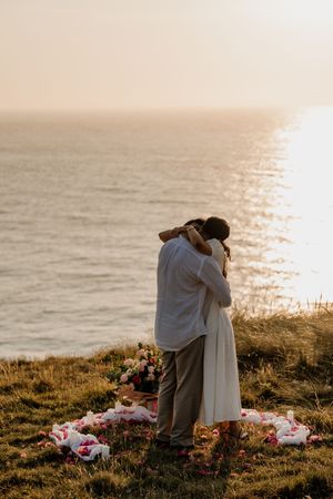 Couple embracing at sunset on a coastal cliff with flower petals and bouquet, romantic proposal scene by the ocean.