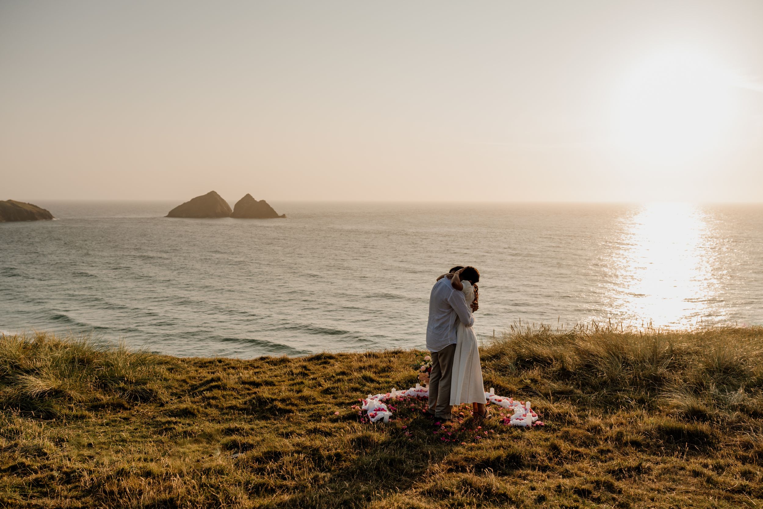 Couple embracing at golden hour on coastal cliffs overlooking the ocean with rocky islands in the background.