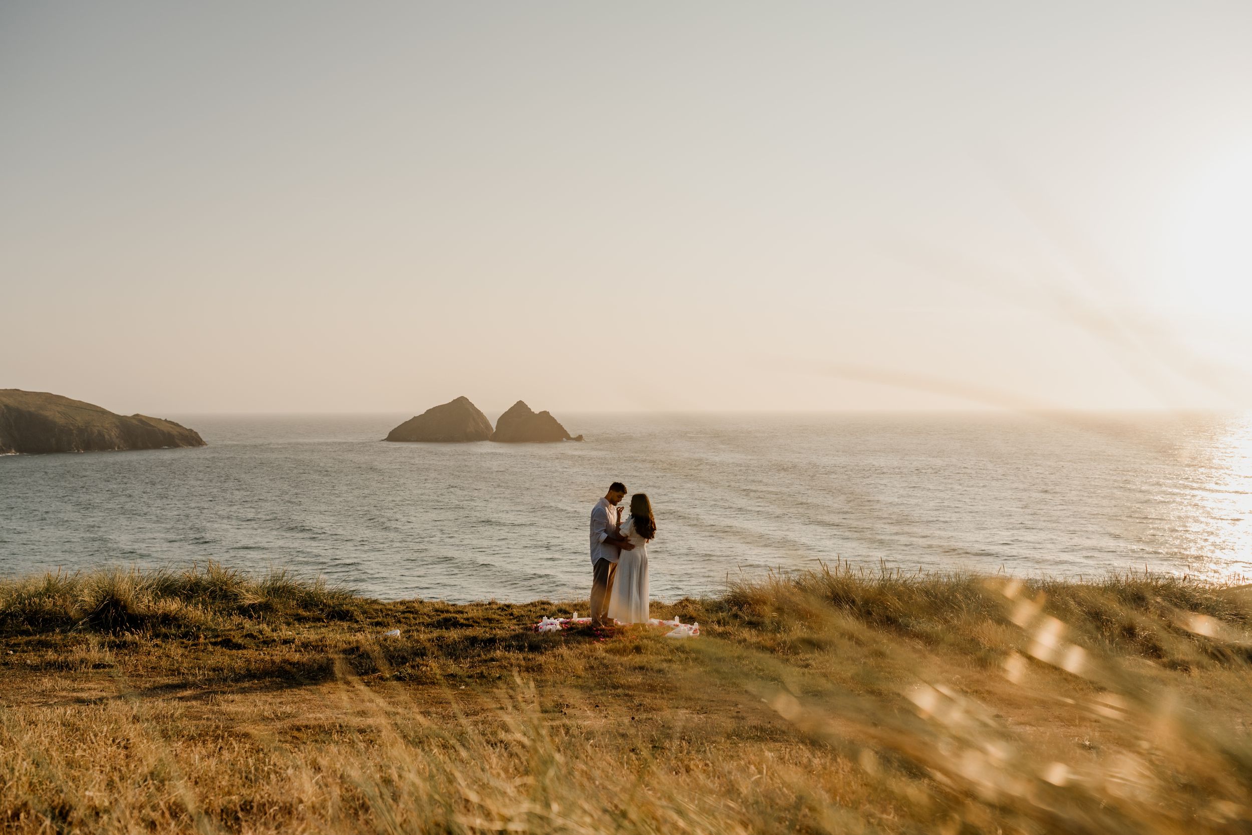 Couple embracing at golden hour on coastal cliffs overlooking rocky ocean islands in warm sunset light.
